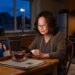 A cinematic, hyper-realistic photograph of a dim kitchen at 3 a.m. A woman in her sixties sits alone at a small wooden table, her face softly lit by the warm glow of a smartphone held in both hands, colorful slot reels faintly reflected in her reading glasses. Beside her: a cooling cup of tea with a spoon resting on the saucer, a folded newspaper, and an old framed photo of a smiling couple propped against the fruit bowl. Through the window behind her, the deep blue-black of a sleeping suburban neighborhood, a single streetlight visible in the distance. The fridge light spills a soft yellow wedge across the linoleum floor. The mood is quiet, intimate, and melancholic — less about gambling and more about solitude, ritual, and memory.